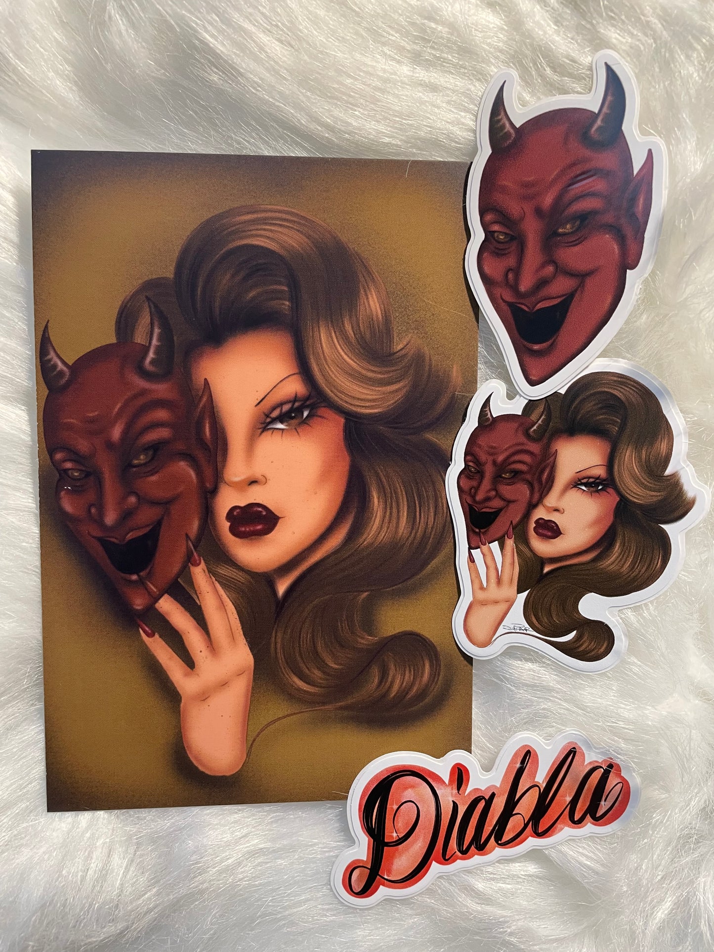 A stylized illustrated print or poster showing a woman with voluminous brown hair, dark red lips, and a red devil mask, displayed on a white faux-fur surface with matching die-cut sticker-style graphics and the word 'Diablo' in red script.